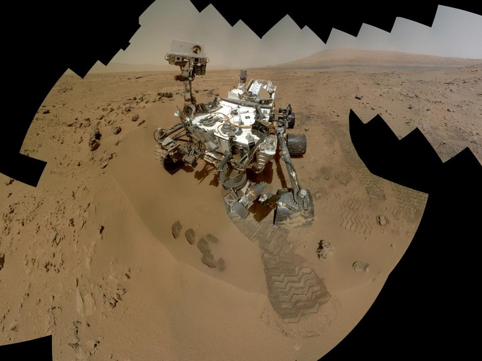 NASA’s Curiosity rover scoops up rock samples on Mars. The planet’s red color comes from rusty iron minerals in the sand and boulders on its surface. (Photo via NASA.gov)